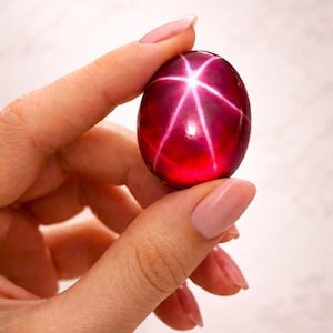 May include: A polished, oval-shaped cabochon gemstone with a deep, translucent red color. The stone features a distinct six-rayed star pattern, known as asterism, which appears as bright white lines intersecting at the center of the surface.