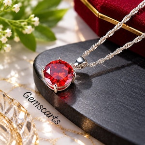 Può includere: Una collana in argento con un ciondolo in pietra preziosa rossa vibrante, rotonda e sfaccettata, montata su quattro griffe e sospesa a una catena d'argento intrecciata. La parola Gemscarts è stampata in nero vicino al ciondolo.