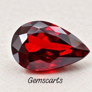 Puede incluir: Una piedra preciosa roja tallada en forma de pera con un aspecto brillante y transparente. La piedra presenta múltiples cortes geométricos que reflejan la luz, creando profundos reflejos carmesí. El texto Gemscarts está impreso en negro debajo de la piedra.