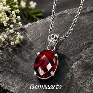 Può includere: Una collana in argento con un ciondolo in pietra preziosa rosso intenso dal taglio ovale e superficie sfaccettata, tenuto da quattro griffe in una montatura in argento decorativa. Il testo Gemscarts è stampato in bianco in basso.