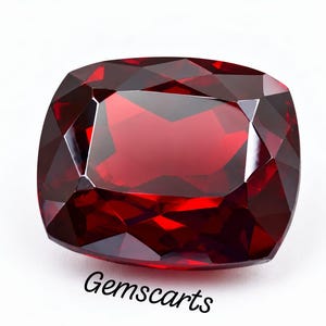 May include: A deep red, cushion-cut gemstone with multiple facets that reflect light, creating a brilliant appearance. The stone has a square shape with rounded corners and is positioned above the black text Gemscarts on a white background.