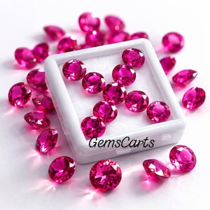 Può includere: Una collezione di pietre preziose rosa, rotonde e sfaccettate, sparse attorno e all'interno di una scatola espositiva quadrata in plastica bianca. La scatola presenta un inserto in schiuma bianca e il testo nero GemsCarts stampato sul bordo.