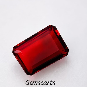 Puede incluir: Una gema de color rojo intenso y vibrante tallada en forma de esmeralda rectangular con bordes facetados. La piedra muestra una gran claridad y un rico tono carmesí. Debajo de la gema, el texto Gemscarts está impreso en una fuente negra simple.