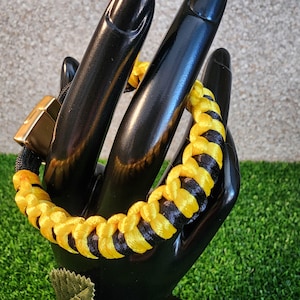 May include: A braided bracelet featuring alternating yellow and black nylon cord segments. The bracelet includes a rectangular metallic clasp with a smooth, reflective finish, securing the woven cord design in a continuous circular loop.