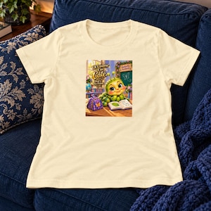 May include: A cream-colored short-sleeve t-shirt featuring a graphic of a green caterpillar with large eyes sitting at a desk with a purple backpack and an open book. The text reads, Of Course I'm Cute, That's the Point, and Butterfly Academy.