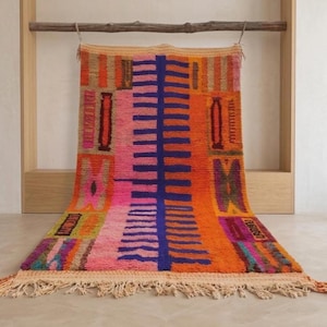 Op de afbeelding: Een rechthoekig vloerkleed met een levendig, veelkleurig geometrisch patroon met een centrale donkerblauwe verticale ruggengraat, geflankeerd door felroze en oranje secties, geaccentueerd met kleine rechthoekige motieven in paars, geel en bruin, afgewerkt met franjes.