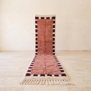 May include: A long, rectangular runner rug featuring a solid dusty rose center with a high-pile texture. The rug is framed by a border of alternating dark brown and cream-colored squares, finished with cream-colored fringe at both ends.