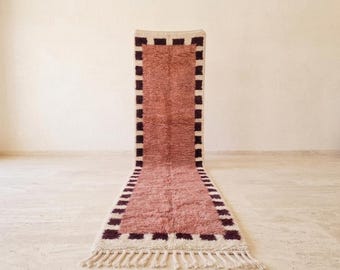 Op de afbeelding: Een lang, rechthoekig loperkleed met een effen, oudroze middenstuk met een hoogpolige textuur. Het kleed is omlijst door een rand van afwisselend donkerbruine en crèmekleurige vierkanten, afgewerkt met crèmekleurige franjes aan beide uiteinden.