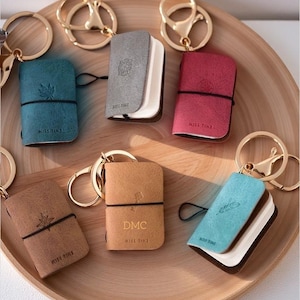 Op de afbeelding: Een verzameling van zes miniatuur notitieboek sleutelhangers in groenblauw, grijs, rood, beige, bruin en lichtblauw. Elke sleutelhanger heeft een goudkleurige metalen ring, een zachte kaft met reliëf iconen en de tekst MISS TIME op de voorzijde.