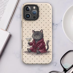 May include: A beige smartphone case featuring a black polka dot pattern and a central illustration of a grumpy grey cat wearing a dark red bathrobe while holding a martini glass with an olive on a toothpick, set against a light background.
