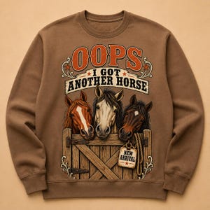 Puede incluir: Una sudadera marrón de cuello redondo con un gráfico de tres caballos mirando por encima de una puerta de establo de madera. El texto dice OOPS I GOT ANOTHER HORSE y NEW ARRIVAL en una etiqueta colgante, con detalles en naranja y blanco.