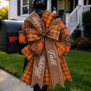 May include: A decorative bow attached to a black mailbox, featuring brown and orange plaid ribbon, brown ribbon with white script text reading Happy Fall, and brown ribbon with orange maple leaf patterns and white swirling line designs.