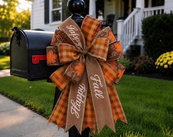 Op de afbeelding: Een decoratieve strik bevestigd aan een zwarte brievenbus, voorzien van bruin en oranje geruit lint, bruin lint met witte tekst Happy Fall, en bruin lint met oranje esdoornbladpatronen en witte krullende lijnontwerpen.