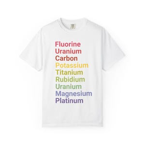Puede incluir: Una camiseta blanca de manga corta con una lista vertical de elementos químicos impresa en un degradado de colores arcoíris. El texto dice Fluorine, Uranium, Carbon, Potassium, Titanium, Rubidium, Uranium, Magnesium y Platinum en orden.
