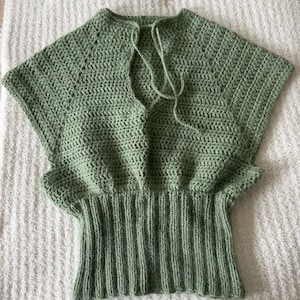 May include: A sage green crochet top featuring short sleeves, a deep V-neckline with thin tie closures, and a wide, vertically ribbed hem. The garment is constructed with a consistent stitch pattern throughout the bodice and sleeves.
