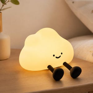 Könnte beinhalten: Ein wolkenförmiges Nachtlicht, das ein warmes, sanftes Licht ausstrahlt. Die Lampe hat eine glatte, cremeweiße Oberfläche mit einem aufgedruckten schwarzen Gesicht, bestehend aus zwei kleinen Punkten als Augen und einem geschwungenen Mund. Zwei schwarze Beine.
