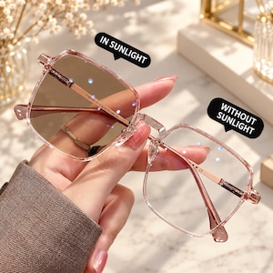 Pode incluir: Óculos fotocromáticos com armação rosa transparente e lentes quadradas. Uma lente está tingida de castanho com a etiqueta IN SUNLIGHT, a outra é clara com a etiqueta WITHOUT SUNLIGHT. As hastes metálicas trazem o texto TR8008 C15 53-15-146 CE.
