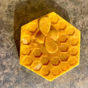 May include: A hexagonal yellow soap bar featuring a raised honeycomb pattern across the surface and a three-dimensional bee figure centered on top. The soap has a textured, speckled appearance with varying shades of golden yellow and orange.
