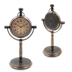 May include: A brass-toned metal desk clock featuring a circular face with dual 12 and 24-hour markings, mounted on a swivel frame with a pedestal base. The reverse side of the clock and the base display an intricate, dark, embossed floral pattern.