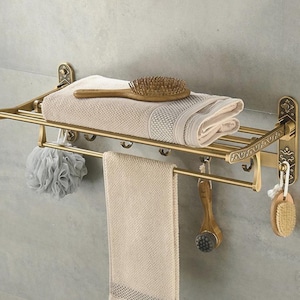 Puede incluir: Un toallero de pared de latón antiguo con estante superior, barra inferior y varios ganchos. Sostiene una toalla beige doblada, un cepillo de madera para el cabello, una esponja de baño gris, un cepillo facial y un pequeño cepillo redondo.