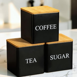 Może przedstawiać: Zestaw trzech kwadratowych czarnych pojemników z jasnobrązowymi drewnianymi pokrywkami. Każdy pojemnik ma z przodu biały napis: COFFEE, TEA oraz SUGAR. Pojemniki są ułożone w stos, jeden na górze i dwa na dole.