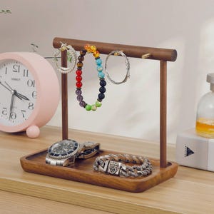 Könnte beinhalten: Ein Schmuckständer aus Holz mit einer horizontalen Stange und einer rechteckigen Ablageschale. An der Stange hängen zwei silberne Creolen und ein buntes Perlenarmband. In der Schale liegen eine silberfarbene Armbanduhr und ein Gliederarmband.