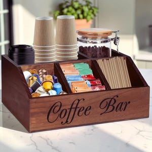 May include: A dark brown wooden coffee bar organizer with the text Coffee Bar printed on the front. It features multiple compartments holding stacks of ribbed paper cups, black lids, coffee pods, sugar packets, wooden stirrers, and a glass jar.