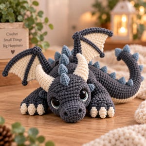 May include: A dark grey crocheted dragon toy featuring cream-coloured horns, wing membranes, and claws. The creature has light blue spikes along its back and tail, large dark eyes, and a textured yarn construction with a soft, rounded appearance.