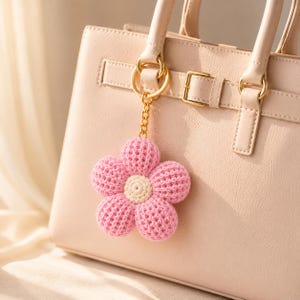 May include: A light pink and cream crocheted flower keyring attached to a gold-toned metal ring and chain, hanging from the front of a structured, light beige handbag featuring a horizontal strap detail with a gold-toned metal buckle.