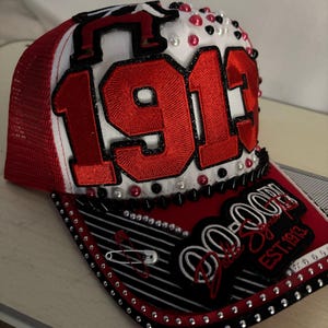May include: A red and white trucker hat featuring the number 1913 in large red embroidered numerals on the front panel. The hat is decorated with red, white, and black beads, silver studs, a safety pin, and text reading Delta Sigma EST. 1913.