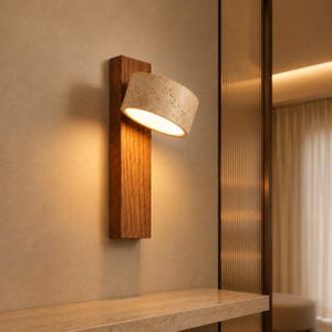 Op de afbeelding: Een wandlamp met een rechthoekige basis van donker hout en een cilindervormige, getextureerde beige stenen kap. De lamp verspreidt een warme, neerwaartse gloed die de natuurlijke houtnerf en het poreuze oppervlak van de steen benadrukt.