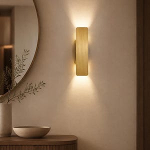 May include: A rectangular wall sconce with a brushed brass finish, emitting a warm, dual-directional light beam that illuminates the wall above and below the fixture, creating a soft glow on the surrounding neutral-toned wall surface.