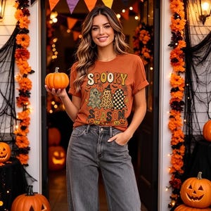 Peut inclure: Un t-shirt à manches courtes orange brûlé avec le texte "SPOOKY SEASON" dans une police rétro, accompagné de trois illustrations de fantômes aux motifs camouflage, léopard et à carreaux, porté avec un jean en denim gris anthracite.