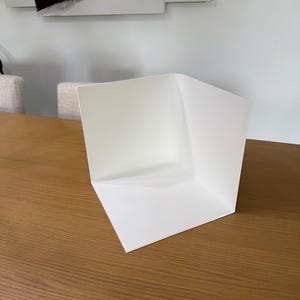 Op de afbeelding: Een wit L-vormig displaybord bestaande uit twee loodrechte panelen die verbonden zijn bij een centrale verticale naad. De structuur is gemaakt van een glad, mat materiaal en vormt een strakke hoek die dient als een neutrale achtergrond.