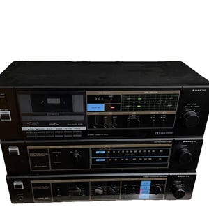 May include: A black Sanyo stereo system consisting of a cassette deck, an AM/FM tuner, and an integrated amplifier stacked vertically. Each unit features various control knobs, push buttons, and illuminated displays with text labels in white.