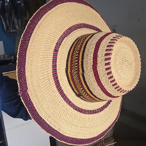 Puede incluir: Un sombrero tejido de ala ancha con una base de paja de color tostado natural, decorado con patrones circulares concéntricos en burdeos intenso, morado oscuro y amarillo mostaza. El sombrero tiene una copa plana y un ala circular ancha.