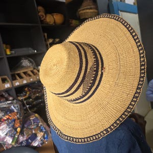 May include: A wide-brimmed woven straw hat featuring a natural tan colour with dark navy blue circular bands around the crown and a matching intricate geometric pattern along the outer edge of the brim, constructed from tightly woven plant fibres.