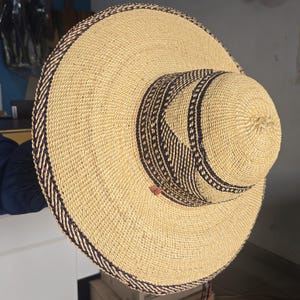 May include: A wide-brimmed straw hat featuring a natural tan woven texture with dark brown braided trim detailing along the edge of the brim and around the base of the crown. The hat has a rounded top and a wide, flat brim with a small red accent.