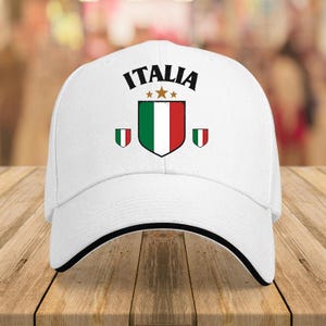 Puede incluir: Una gorra de béisbol blanca con la palabra ITALIA en texto negro sobre un escudo con los colores de la bandera italiana: verde, blanco y rojo. Tres pequeñas estrellas doradas se sitúan sobre el escudo, flanqueadas por dos escudos menores.