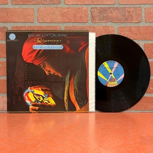 May include: An Electric Light Orchestra "Discovery" vinyl record album. The album cover features a person in a red head covering, with the album title and band name. The vinyl record is black with a colorful label.