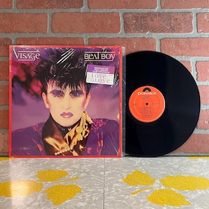 May include: A Visage Beat Boy vinyl record album with a purple and black cover featuring a person with dark hair and purple makeup. A white sticker reads Contains the full-length club version of Love Glove. A black vinyl disc with an orange label.
