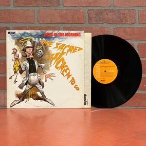 May include: An album cover for "Guys in the Morning" featuring a cartoon illustration of a man holding a chicken. The album is titled "One Sacred Chicken to Go". A black vinyl record is next to the album cover.