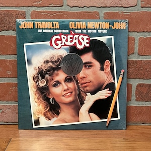 May include: A vinyl record of the Grease soundtrack featuring John Travolta and Olivia Newton-John. The album cover has a photo of the two actors. The album is labeled "The Original Soundtrack From The Motion Picture" and includes the text "Grease". A pencil rests on the album.