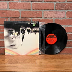 May include: A vinyl record album titled "More Hot Rocks (Big Hits & Fazed Cookies)" by The Rolling Stones. The album cover features a black and white image of the band members with a rainbow at the bottom. The record is black with a red and white label.