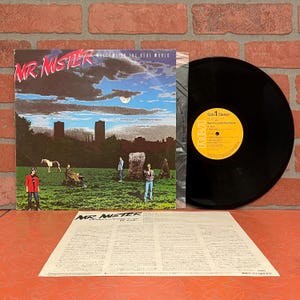 May include: An album cover for "Welcome to the Real World" by Mr. Mister. The cover art features a landscape with figures, a horse, and a cloudy sky. The vinyl record is visible next to the cover, with the album's label showing.