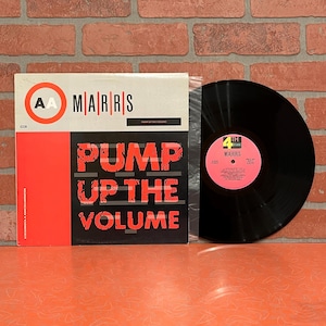 May include: A vinyl record album by MARRS, titled "Pump Up The Volume." The album cover is white, black, and red, with the title in large, bold letters. The record itself is black and in a plastic sleeve. The background is a brick wall.