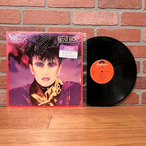 May include: A black vinyl record with a red and orange label that says "Polydor" and "Visage Beat Boy". The record is in a plastic sleeve with a red and orange album cover featuring a man with black hair and makeup. The album cover says "Visage" and "Beat Boy" and "Contains the 12" inch club version of Love Glove".