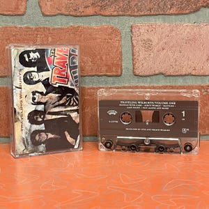 May include: Two clear cassette tapes. The first cassette has a cover with the text "TRAVELING WILBURYS" and a photo of the band. The second cassette tape has the text "TRAVELING WILBURYS/VOLUME ONE" and song titles.