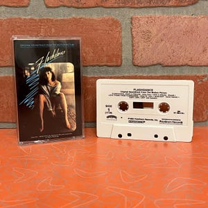 May include: A vintage cassette tape of the "Flashdance" soundtrack. The cassette case features a woman in a seated pose. The white cassette tape has the title "Flashdance" printed on it.