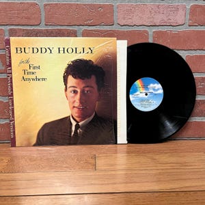 May include: A vinyl record album by Buddy Holly, titled "For the First Time Anywhere." The album cover features a portrait of Buddy Holly. The record is black, with a label showing a rainbow and the MCA Records logo.